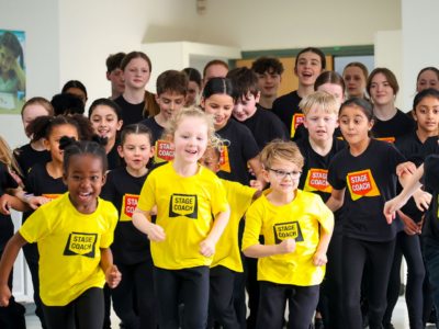A joyful group of Stagecoach students in black and yellow uniforms running toward the camera during a performing arts activity.