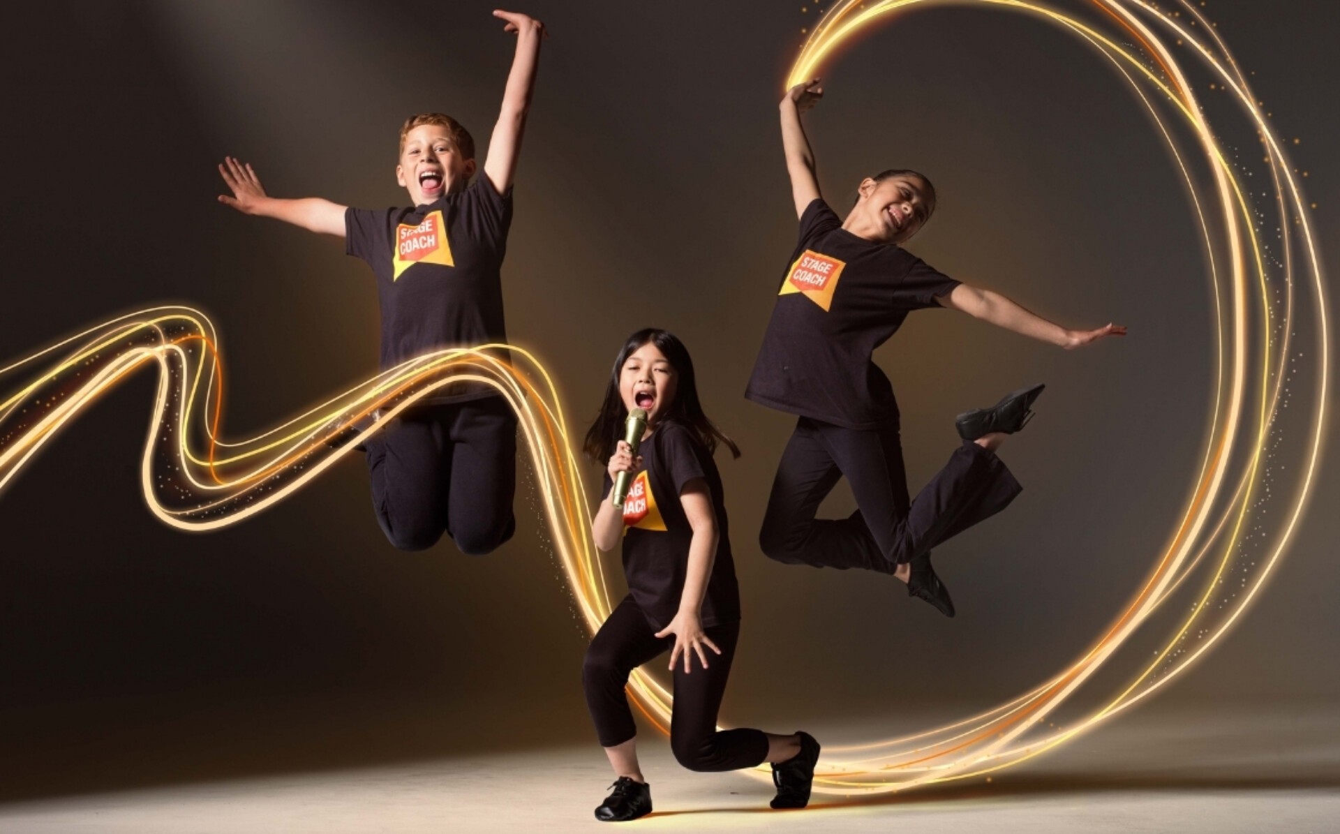 Three young performers in black Stage Coach T-shirts pose energetically on a studio stage, with two jumping and one kneeling with a microphone, surrounded by glowing golden light trails.
